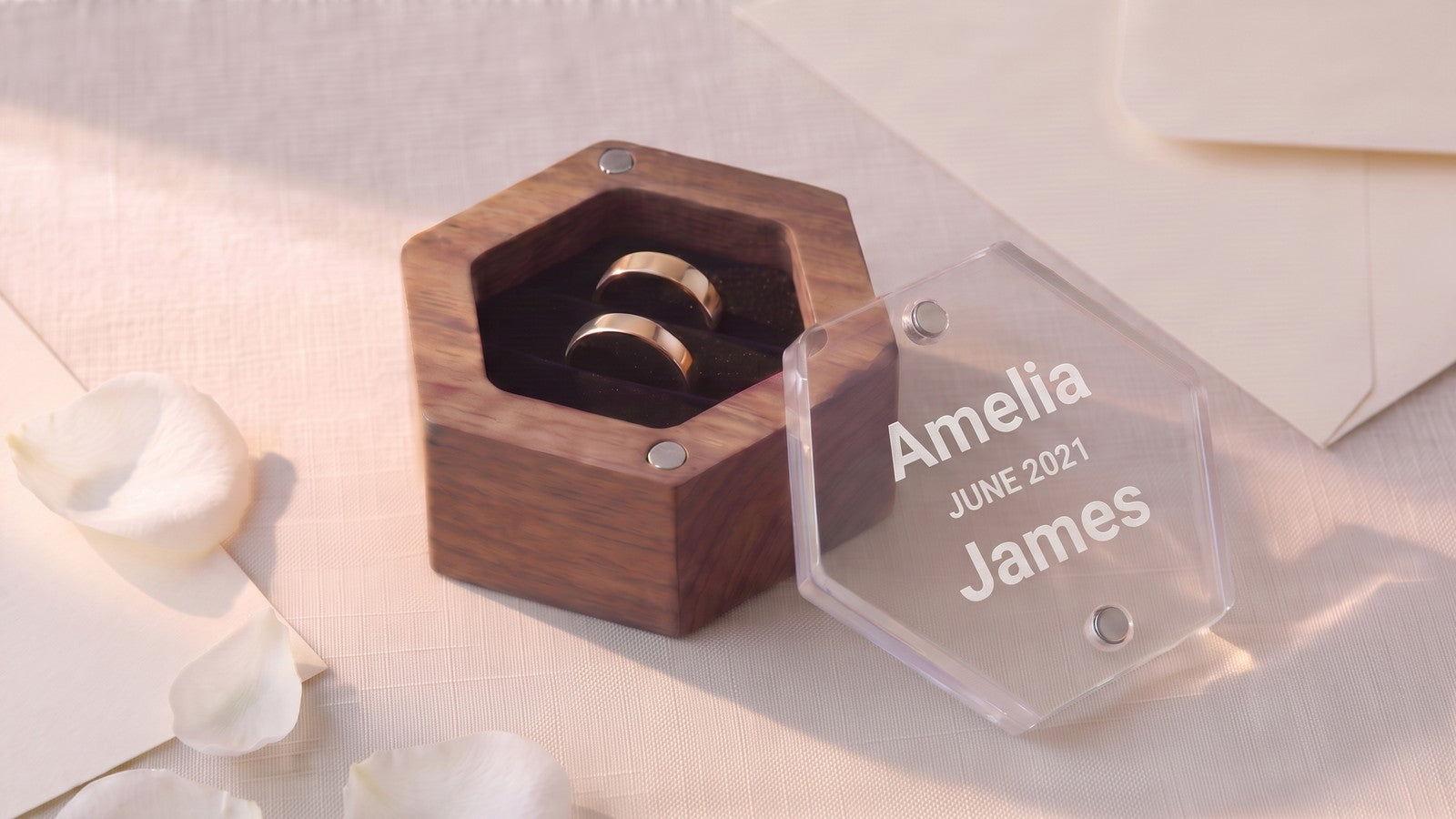 Hexagonal walnut double ring box on wedding morning, clear acrylic lid lifted off beside it and engraved with two names and a date, one gold band in each of the two velvet rows